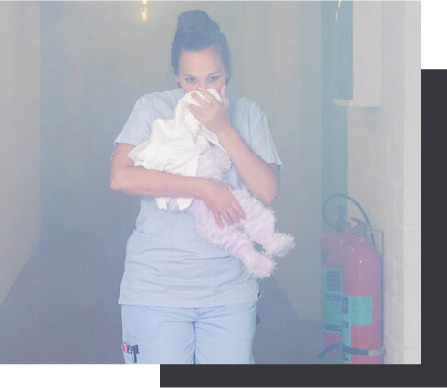 Photo of a nurse carrying a baby and covering her nose walking out of a smoke-filled hospital hallway. Black box in the background.