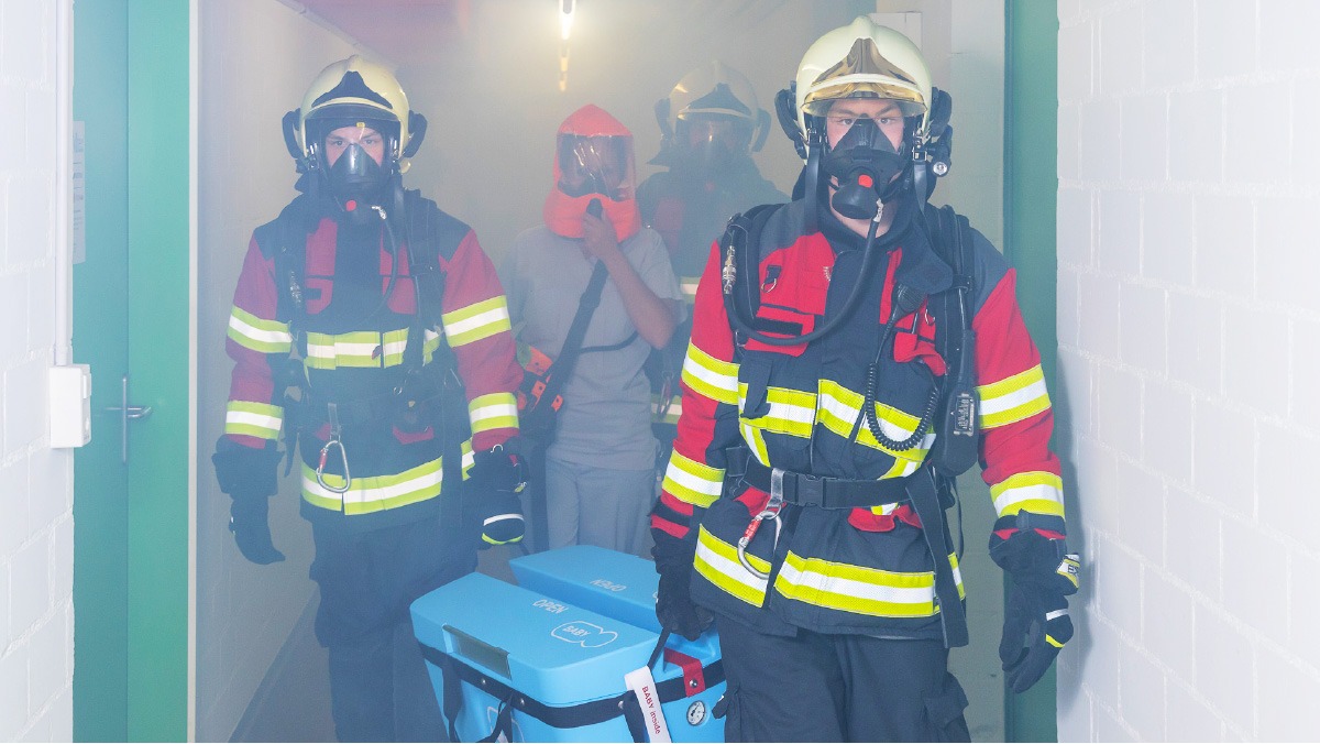 Three fully equipped firemen and a nurse with a breathing device carrying out a Lifebox device from a smoke-filled hospital hallway.